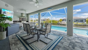 A covered outdoor area with a pool, dining table, chairs, and a kitchen area. - BRAND NEW - Pool overlooking canal, Outdoor Grilling station - Villa Hummingbird House - Roelens (Cape Coral)