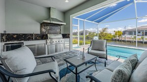 A modern outdoor kitchen with a grill, dining area, and a view of a swimming pool. - BRAND NEW - Pool overlooking canal, Outdoor Grilling station - Villa Hummingbird House - Roelens (Cape Coral)