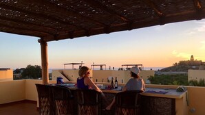 Une terrasse sur le toit comprenant un bar, des espaces de détente en plein air et une vue sur une colline lointaine où se trouve un bâtiment.