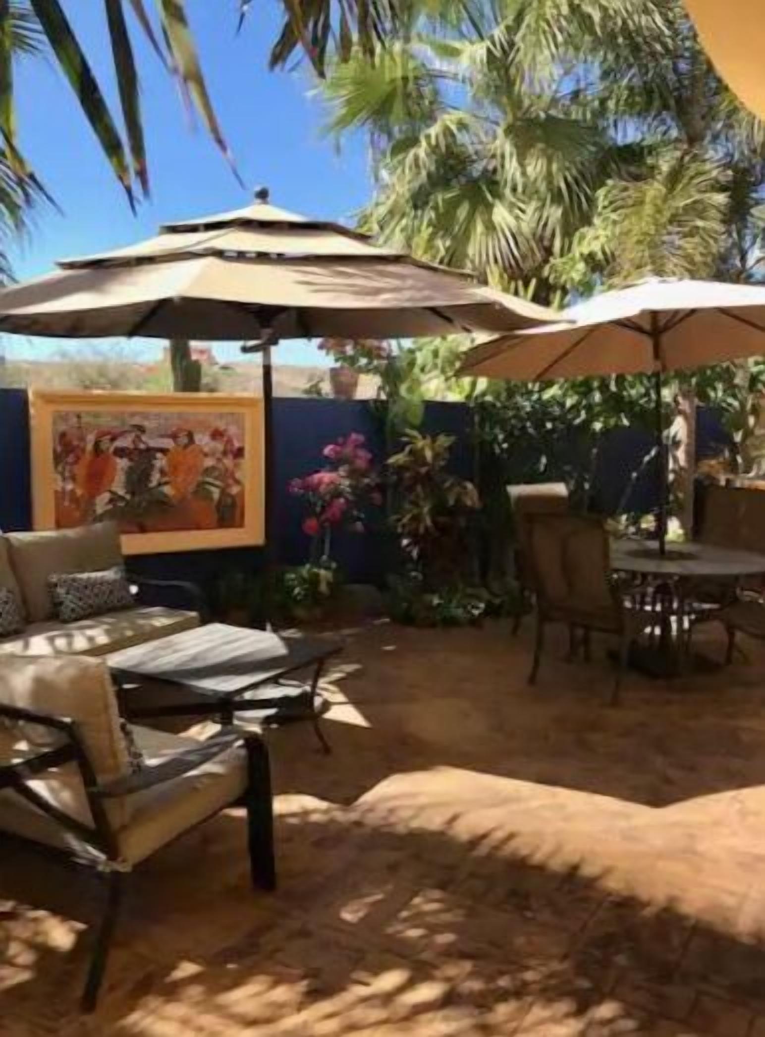 An outdoor patio area with a dining table and chairs, surrounded by palm trees and a mural on the wall.