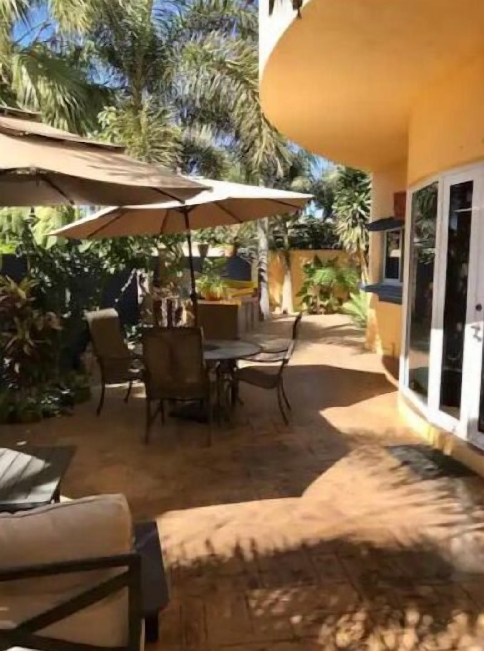 An outdoor patio area with tables and chairs, surrounded by palm trees and covered by umbrellas.