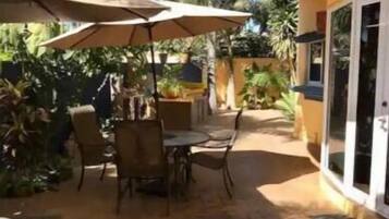 An outdoor patio area with tables and chairs, surrounded by palm trees and covered by umbrellas.