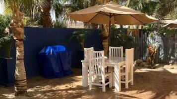 A patio area with a table and chairs, covered by a large umbrella, surrounded by palm trees and a blue wall.