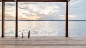Een houten terras met een metalen trap, uitkijkend over een kalm wateroppervlak en een verre kustlijn tijdens de zonsondergang.