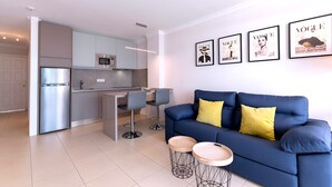 A modern living room with a blue sofa, two round side tables, and a kitchen area with a refrigerator and bar stools.