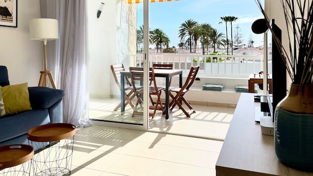 A balcony with a dining table and chairs, a sofa with cushions, a lamp, and a view of palm trees and buildings.