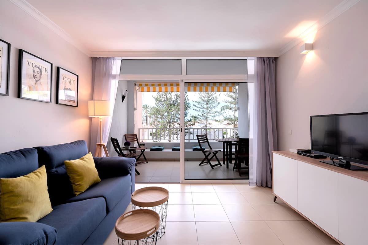 Ein modernes Wohnzimmer mit einem blauen Sofa, einem Fernseher auf einem weißen Schrank und einem Balkon mit Tisch und Stühlen.