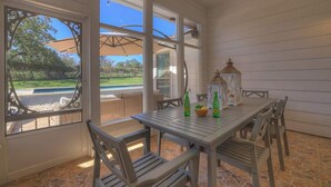 An outdoor patio with a table and chairs, large windows, and a view of a pool and greenery.