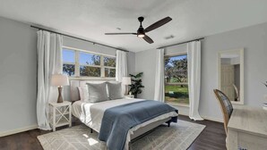A bedroom with a bed, nightstands, a desk, a chair, a ceiling fan, and large windows.