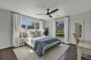 Une chambre à coucher comprenant un lit, des tables de chevet, un bureau, une chaise, un ventilateur de plafond et de grandes fenêtres.