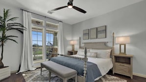 A bedroom with a bed, two bedside tables, a bench, a ceiling fan, and a large window with curtains.