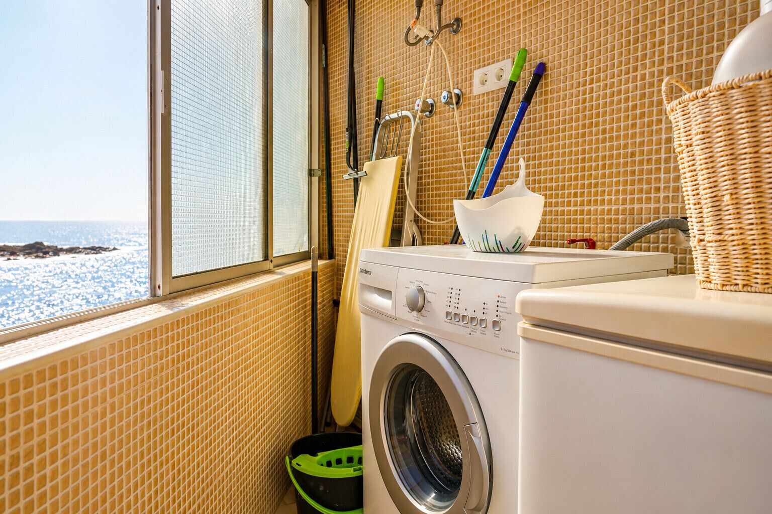 A laundry area with a washing machine, a window with a view of the sea, and various cleaning tools.
