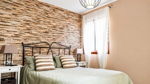 A bedroom with a brick-patterned wall, a bed with green bedding, two bedside tables with lamps, and a window with sheer curtains. - The Waves · Natural Pools & Sea Views, El Pris (El Pris)