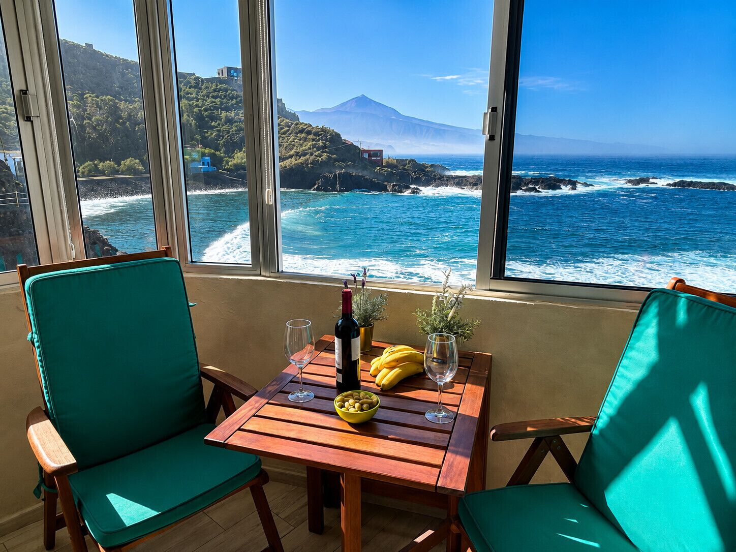 Une table avec une bouteille, un verre, des bananes et un bol d’olives, donnant sur une vue côtière avec des vagues et une montagne.