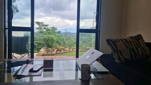 A room with a large window, a laptop on a glass table, a chair, and a couch with a patterned pillow. - 20 min walk to Kandy city from this breezy, quite property with a parking. (Kandy)