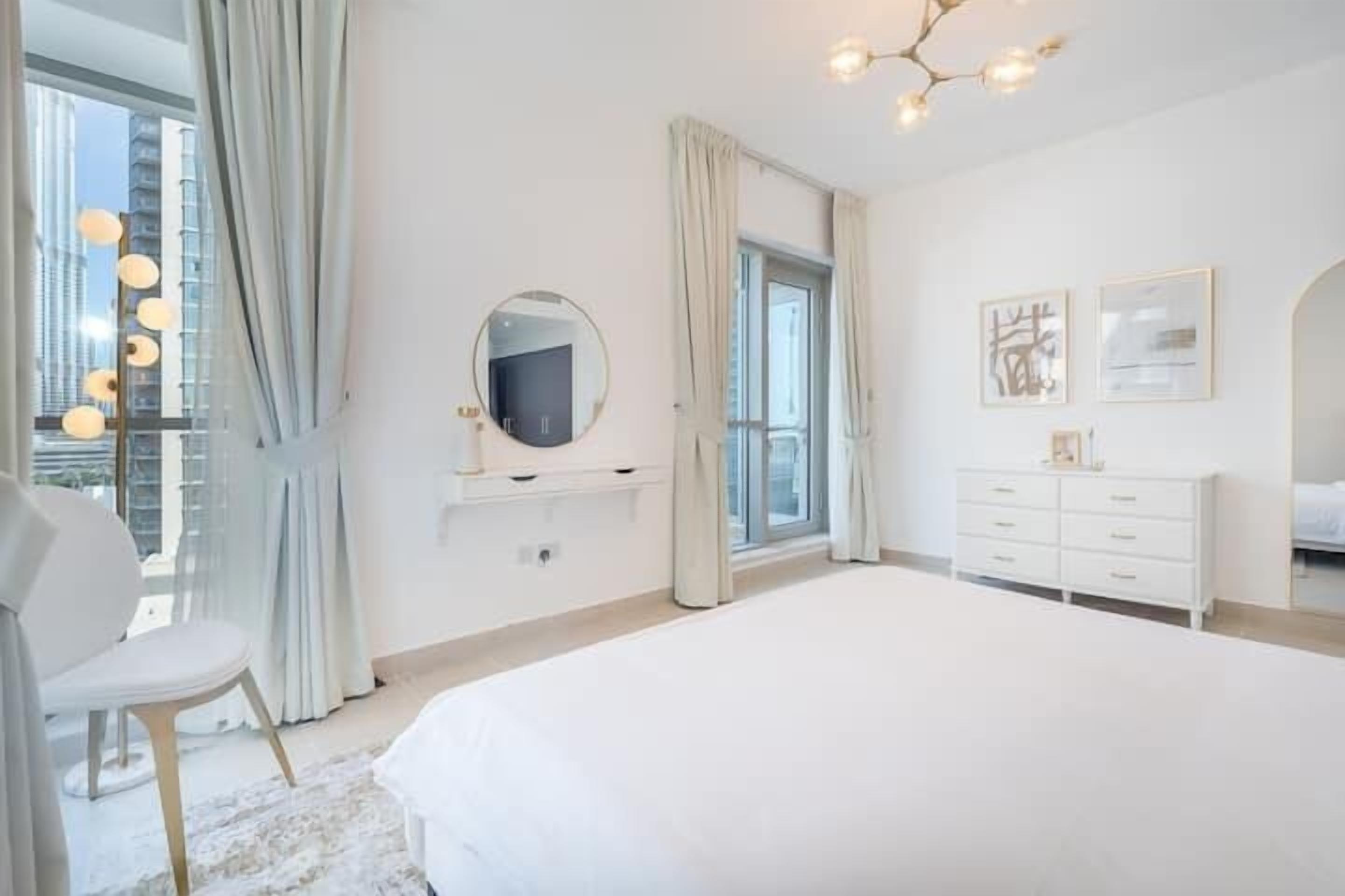 Een moderne slaapkamer met een groot bed, een kaptafel met spiegel en een dressingkast. Er is een raam met gordijnen, en een kroonluchter hangt aan het plafond.