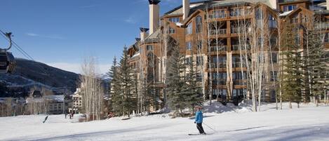 A ski resort with a large lodge featuring multiple chimneys and balconies, surrounded by snow-covered trees and a clear sky.