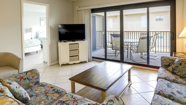 A hotel room with a sofa, a coffee table, a TV on a stand, and a balcony with outdoor furniture. - Sandy Shores Condo Across From Johns Pass! (Madeira Beach)