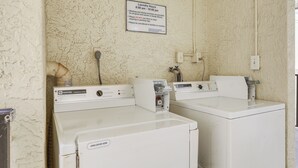 A laundromat with two white washers and dryers, a sign indicating laundry hours, and a utility wall with electrical outlets. - Sandy Shores Condo Across From Johns Pass! (Madeira Beach)