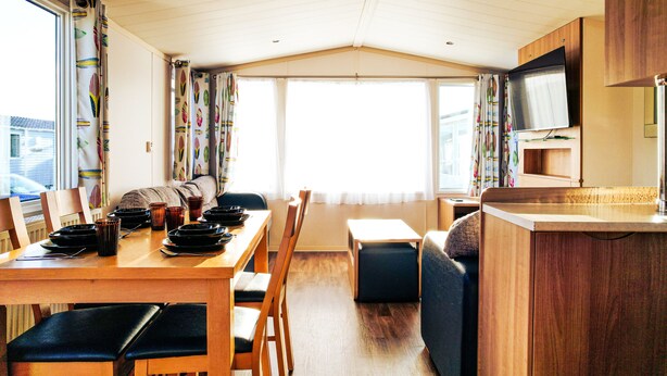 A compact, well-lit room with a dining table, chairs, a sofa, a TV, and a small kitchenette.