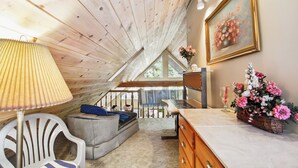 A cozy attic room with a wooden ceiling, a comfortable sofa, a dining table, and a painting on the wall.