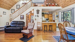 A spacious living area with a black leather sofa, a patterned armchair, and a dining table with wooden chairs. A staircase leads to a loft with a railing and decorative pillows.