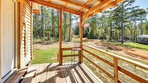 A wooden deck with a metal chair, overlooking a forested area with a dirt path and a vehicle in the background. - Quaint Abbeville Cabin Studio w/ Gas Grill! (Abbeville)
