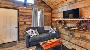 A cozy cabin interior with a wooden ceiling, a gray sofa with patterned cushions, a wooden coffee table, and a mounted flat-screen TV. - Quaint Abbeville Cabin Studio w/ Gas Grill! (Abbeville)
