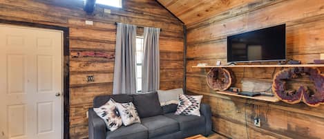 Ein gemütliches Blockhaus-Interieur mit Holzbalkendecke, einem grauen Sofa mit gemusterten Kissen, einem Holztisch und einem an der Wand befestigten Flachbildfernseher.