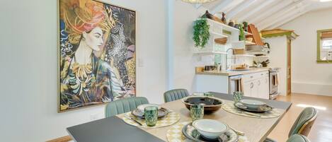 A dining area with a large painting, a modern kitchen in the background, and a dining table set with plates and glasses.