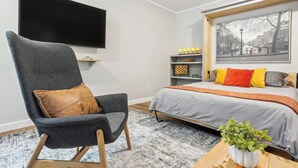 A modern bedroom with a large bed, a grey armchair, a flat-screen TV, and a cityscape artwork. - Pet-friendly Kent Vacation Rental, Walk to Lake! (Covington)