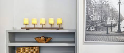 A room with a white wall, a shelf with five wall-mounted lamps, a wicker basket, a polka-dotted stool, and a framed picture of a street scene.