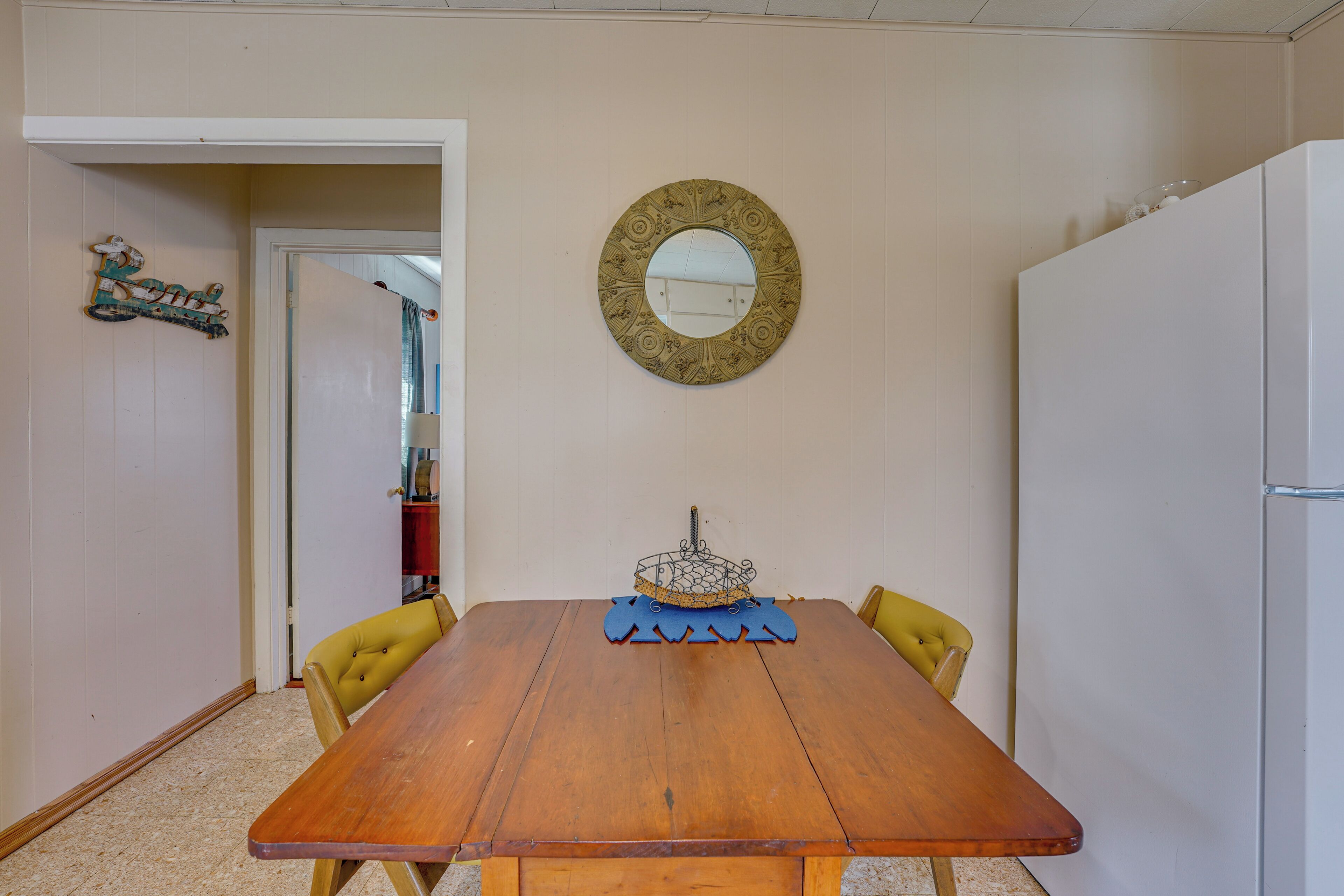 Un comedor con una mesa de madera, dos sillas amarillas, un espejo decorativo y un refrigerador blanco.