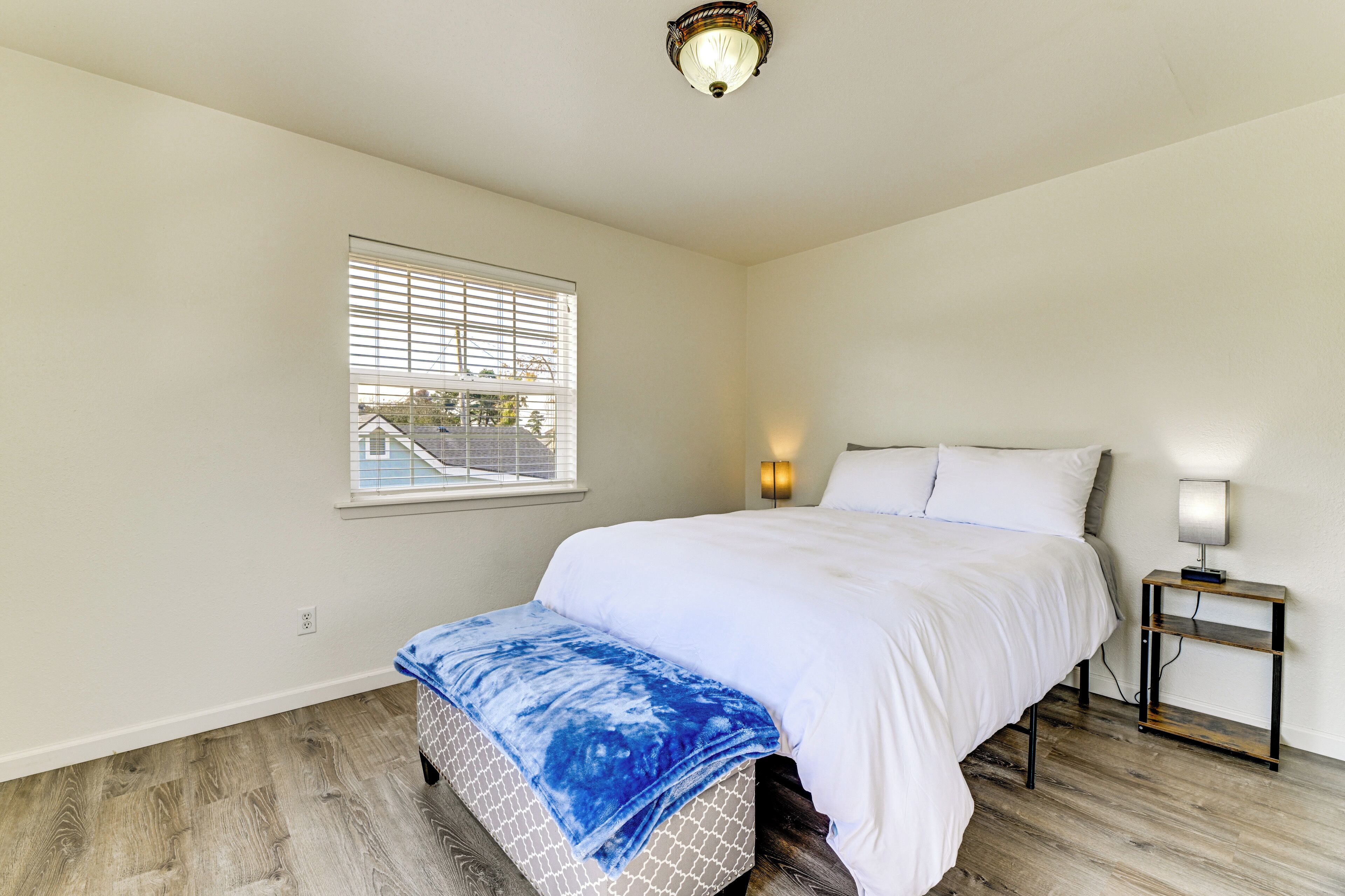 A bedroom with a bed, a nightstand, a lamp, a window with blinds, and a ceiling light fixture.