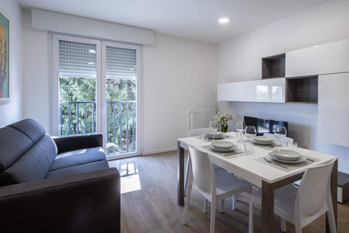 Un comedor moderno con una mesa blanca para cuatro, un sofá gris y un balcón con vistas a zonas verdes.