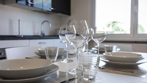 A dining table set with white plates, clear glasses, and a wine glass, with a kitchen in the background.