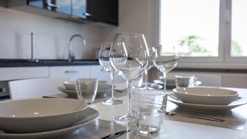 Una mesa de comedor dispuesta con platos blancos, vasos transparentes y una copa de vino, con una cocina al fondo.