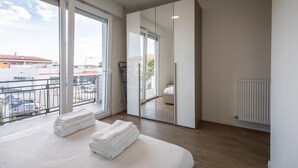 A modern bedroom with a large window, a balcony, and a view of a building with a red roof.