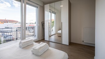 Un dormitorio moderno con un ventanal grande, un balcón y vistas a un edificio de techo rojo.