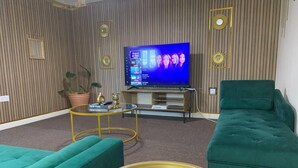 Un salon doté d’un grand téléviseur à écran plat, de deux canapés verts, d’une table basse en verre et d’un panneau décoratif mural.