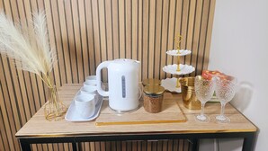 Une table en bois, une bouilloire blanche, un plateau blanc avec des tasses, un plateau doré avec un présentoir blanc, un pot doré et deux verres à vin transparents.