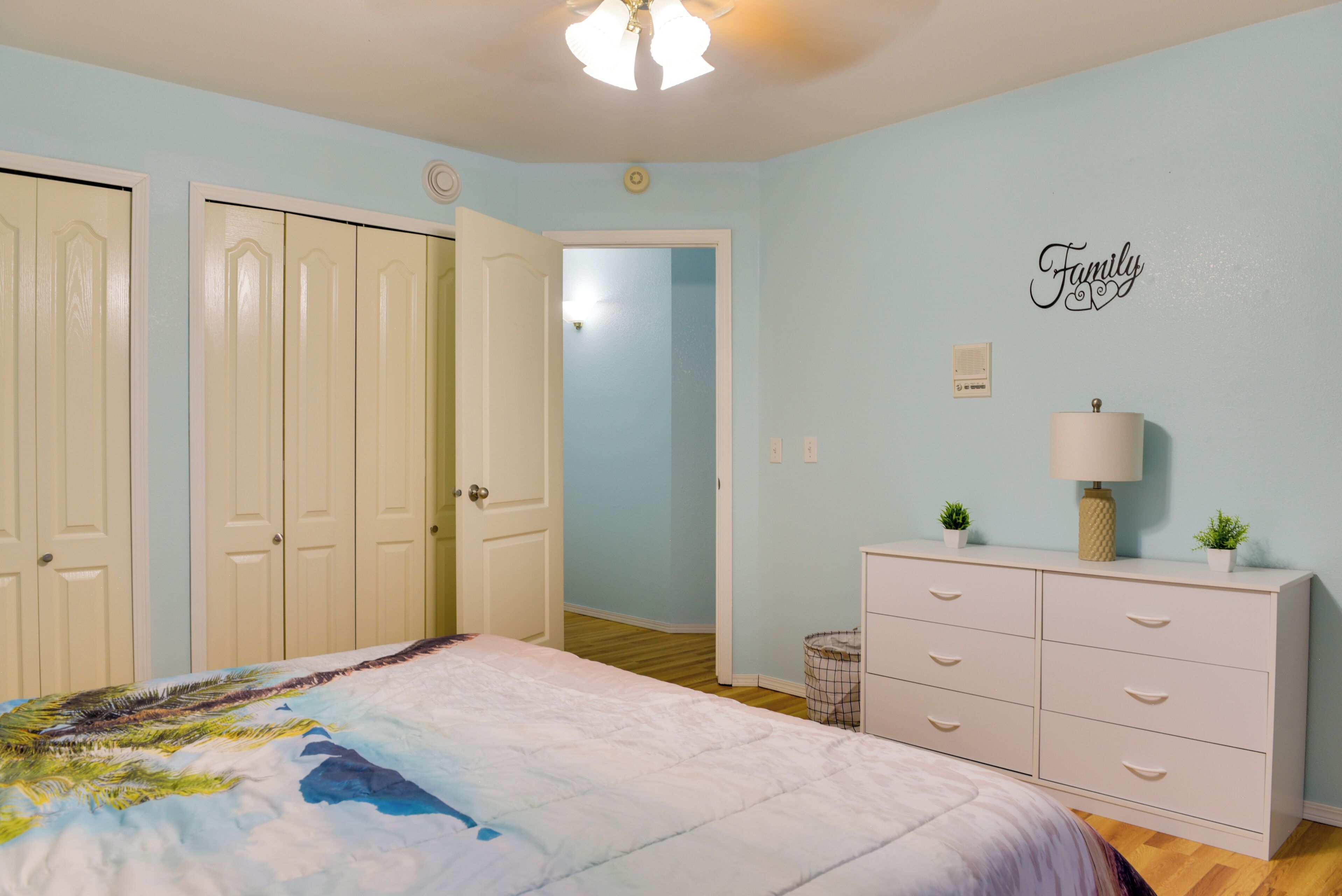 Une chambre à coucher comprenant un lit, une commode, une lampe fixée au mur et une porte donnant sur une autre pièce.