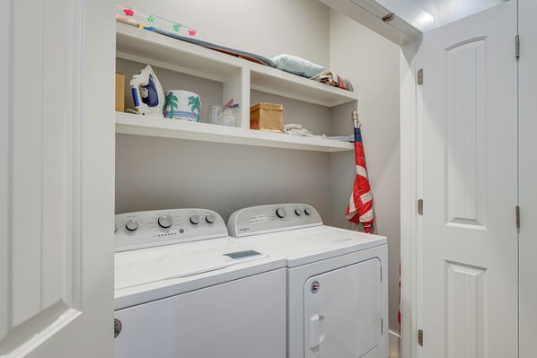 Un cuarto de lavandería con dos lavadoras y secadoras apiladas, repisas superiores con varios artículos y una puerta a la derecha.