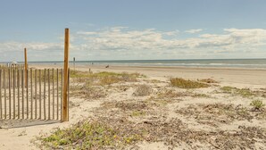 Una playa arenosa con un cerco de madera y vegetación escasa.