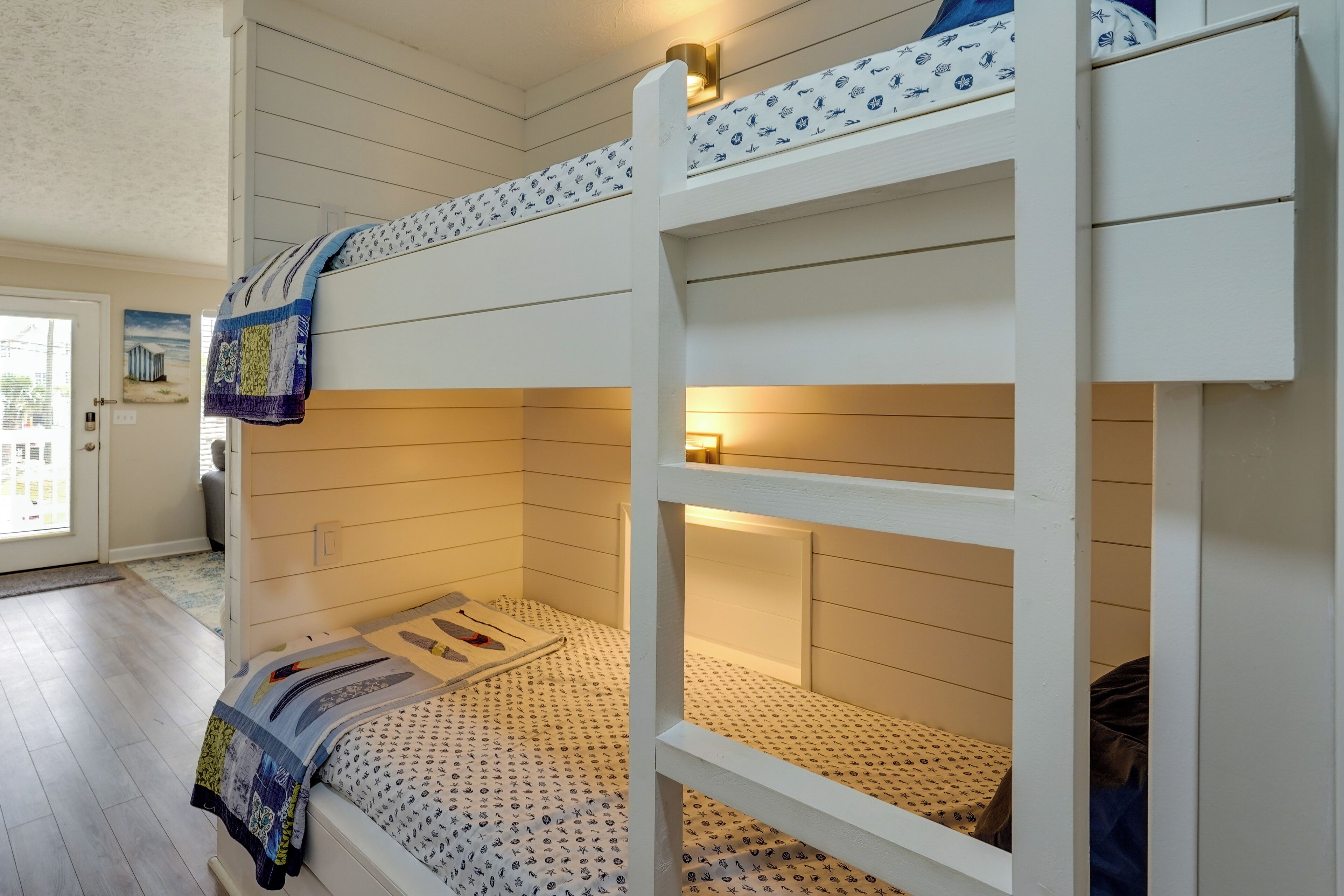 A bunk bed with a patterned bedspread, a small bedside table with a lamp, and a window with a view of the outside.