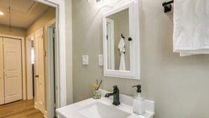 Ein Badezimmer mit einem weißen Waschbecken, einem schwarzen Wasserhahn und einem Spiegel darüber. Ein Handtuch hängt an einem Haken und eine Handseifenflasche steht auf der Ablagefläche.