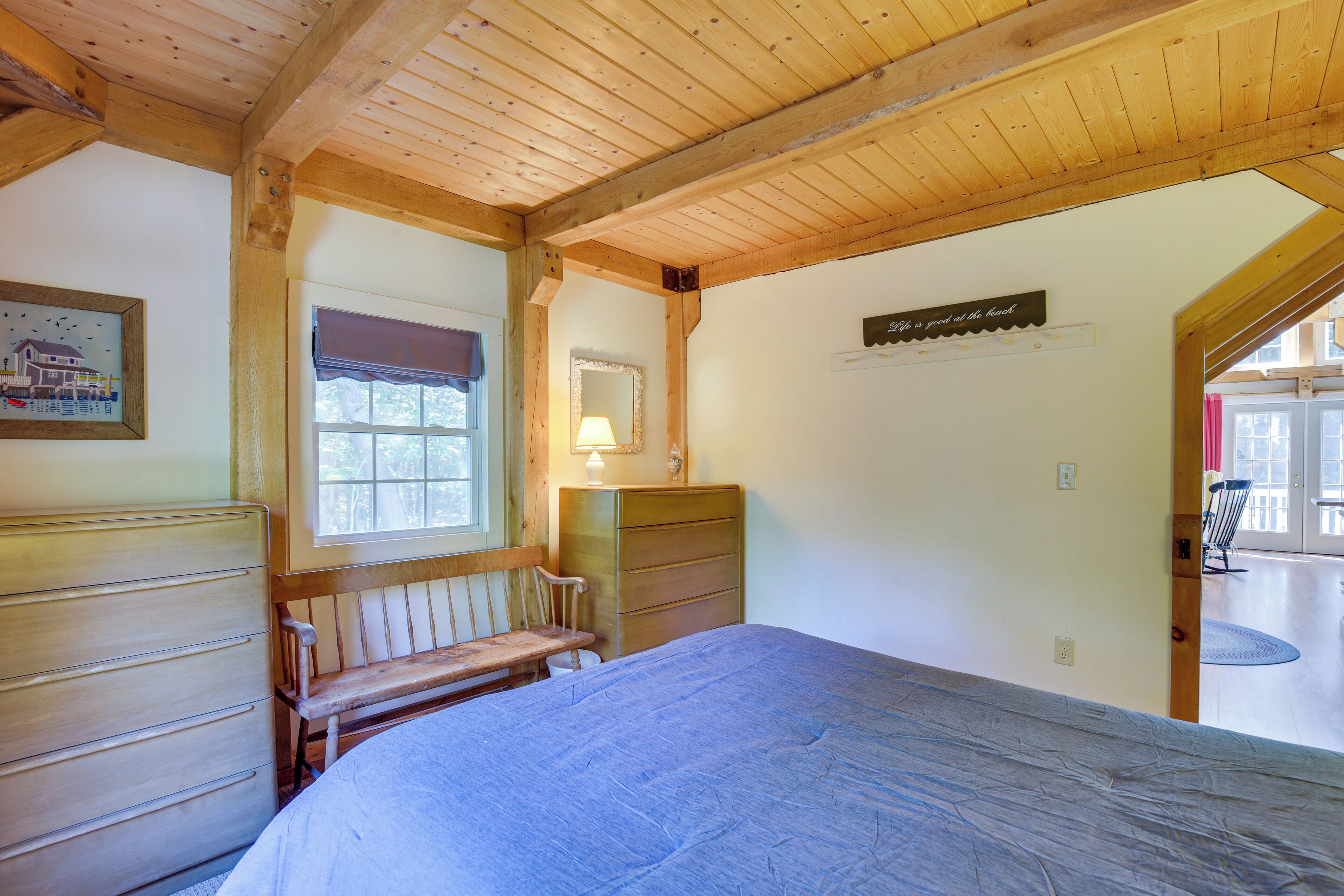 Un dormitorio con techo de madera, cama, cómoda, banca y ventana con vistas, además de un espejo.