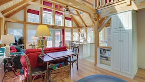 A spacious living room with a high ceiling, wooden beams, a dining area, and a kitchen with white cabinets. - Frye Island Family Cabin With Lake & Beach Access! (Raymond)