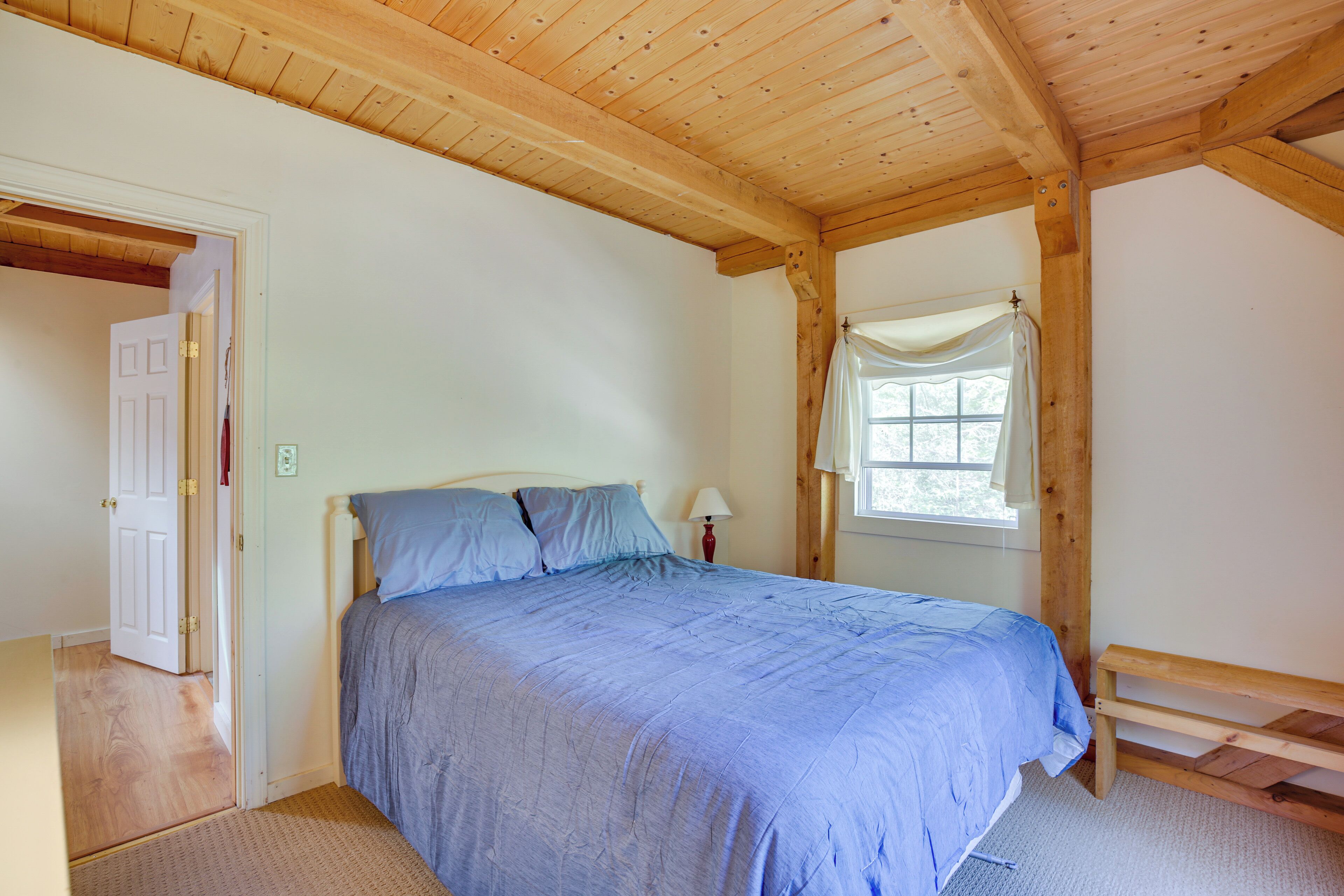 A bedroom with a bed, wooden ceiling beams, a window with a curtain, and a door.