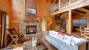 A cozy log cabin interior with a stone fireplace, a gray sofa with animal-themed pillows, a rocking chair, and a spiral staircase.